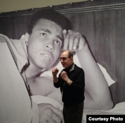 លោក George Kalinsky ជា​អ្នក​ថតរូប​លោក Ali អស់រយៈពេល​ជាង​៥០ឆ្នាំ ឈរ​នៅ​មុខ​ផ្ទាំង​រូបថត​លោក Ali ដ៏​ធំមួយ។ (រូបថ​តផ្តល់​ឲ្យ​ដោយ New York Historical Society)