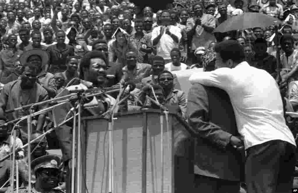 Muhammad Ali embrasse le président du Zaïre Mobutu Sese Seko sur la joue comme le président le présente à la foule à Kinshasa, au Zaïre, le 22 septembre 1974. À gauche, un George Foreman amusé, qui a serré la main du président plus tard. Les deux boxeurs ont assisté aux cérémonies d'ouverture d’un festival au stade du 30 mai, où ils vont se battre pour le titre des poids lourds le 30 octobre 1974. (AP Photo / Horst Faas)