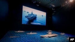 A TV screen showing the U.S. Navy fleet sail in formation near the models of Liaoning aircraft carrier with navy frigates and submarines on display at the military museum in Beijing, Thursday, Aug. 1, 2019. U.S. Secretary of State Mike Pompeo said…